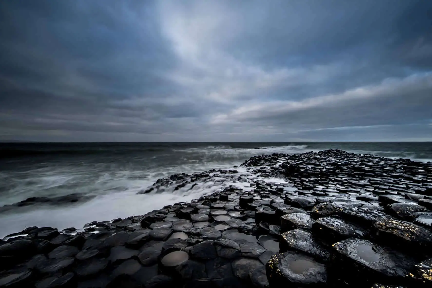 Atlantic waves breaking over dark basalt columns beneath a cloudy sky at the Giant’s Causeway